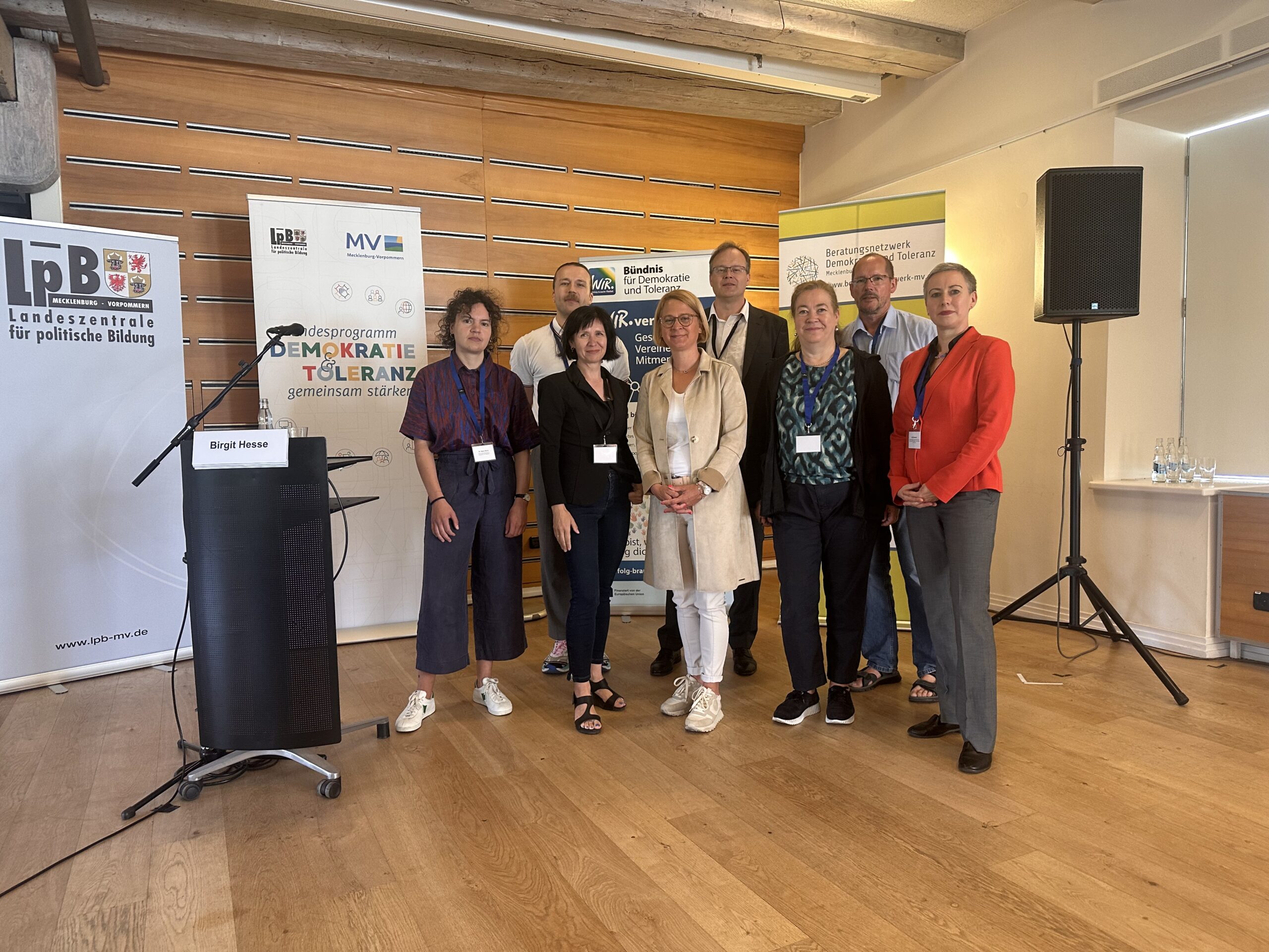 Gruppenfoto von neun Personen vor mehreren Roll-Ups mit den Logos der Landeszentrale für politische Bildung Mecklenburg-Vorpommern, des Landesprogramms „Demokratie und Toleranz gemeinsam stärken!“ sowie des Beratungsnetzwerks Demokratie und Toleranz. Die Teilnehmenden der Sommertagung stehen nebeneinander im Veranstaltungsraum, darunter Vertreterinnen aus Zivilgesellschaft, Politik und Verwaltung. Links im Bild ein Rednerinnenpult mit dem Namensschild „Birgit Hesse“. Alle Personen tragen Namensschilder, der Hintergrund ist geprägt von Holzpaneelen und Bannern der Veranstaltenden.