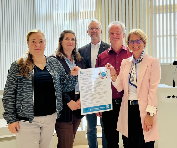 Fünf Personen stehen im Plenarsaal des Landtags Mecklenburg Vorpommern und präsentieren gemeinsam den Wahlaufruf der Initiative „WIR. Erfolg braucht Vielfalt“. Zwei Personen halten das Plakat mit dem Aufruf „WIR gehen wählen“ in die Kamera.